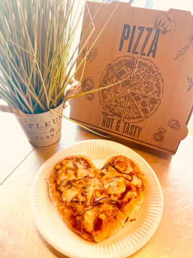 Herzförmige Pizza auf einem Teller, neben einer Pizzaschachtel und einer Pflanze.