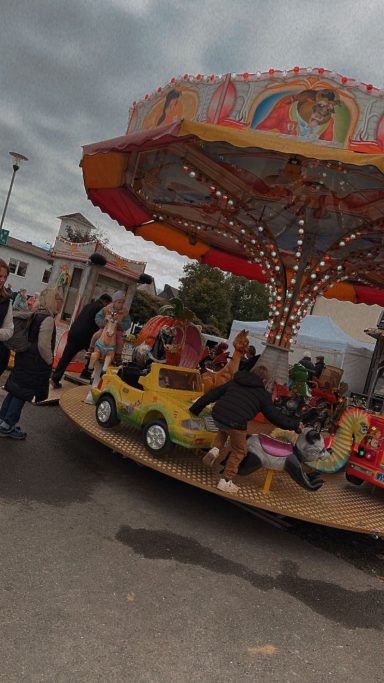 Karussell mit bunten Autos und Kindern, umgeben von Besuchern und unter grauem Himmel.