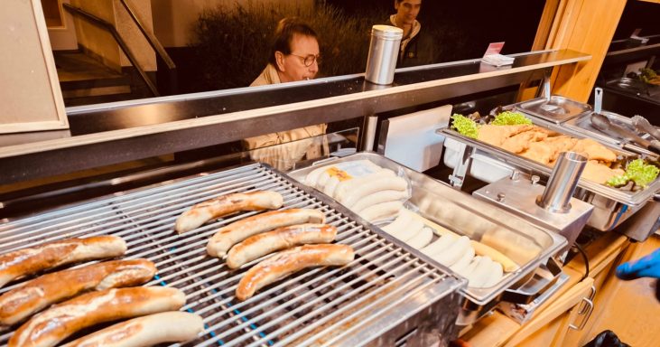 Grill mit Würstchen und einem Verkaufsstand im Hintergrund, frische Beilagen sichtbar.