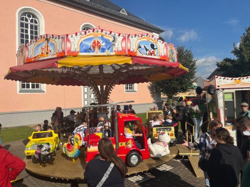 Karussell mit bunten Fahrzeugen und Besuchern auf einem Festplatz.