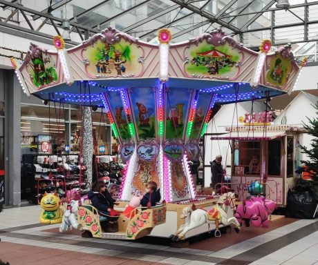 Kettenkarussell mit bunten Lichtern in einem Einkaufszentrum, mehrere Kinder sitzen darin.