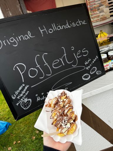 Eine Hand hält eine Portion Poffertjes mit Schokoladensauce und Puderzucker.