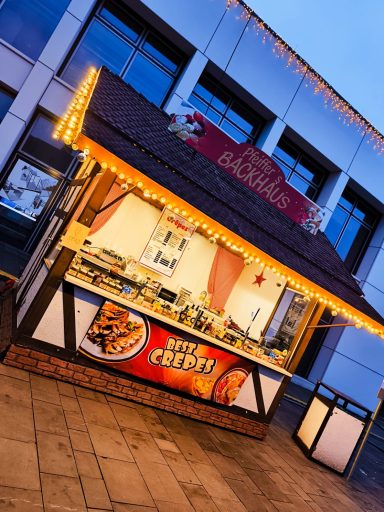 Ein festlich beleuchteter Stand verkauft Crepes und Snacks, umgeben von modernen Gebäuden.