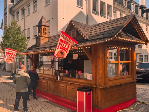 Holzhütte mit Verkaufsstand, rot-weißen Schildern und Besuchern auf einer Straße.