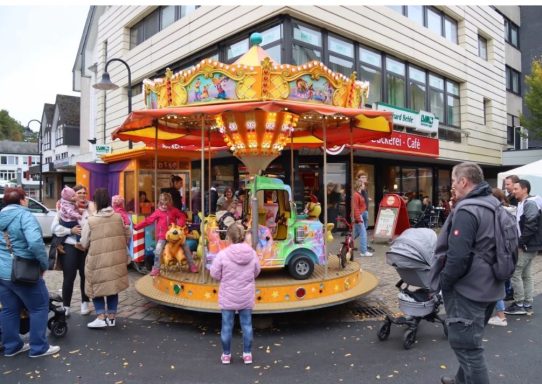 Karussell mit bunten Tieren und Kindern, umgeben von Menschen in einer Stadtstraße.
